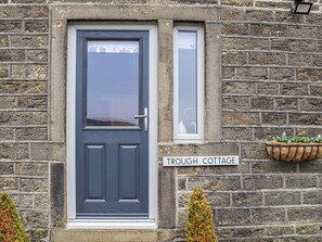 A stone building with a dark grey door, a small window, and a sign reading 'TROUGH COTTAGE.'