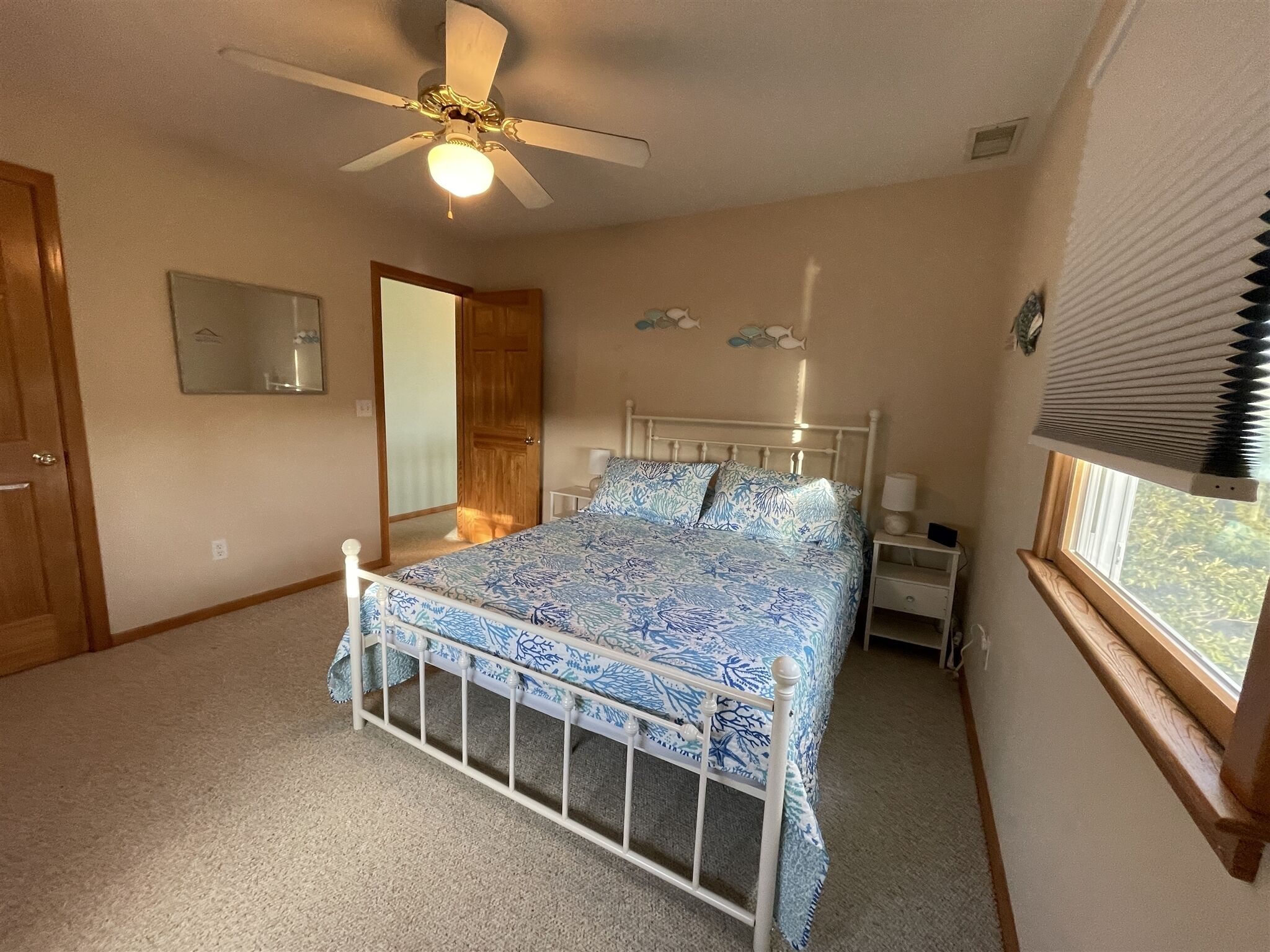 Une chambre à coucher comprenant un lit, un ventilateur de plafond, une table de chevet, une fenêtre avec des stores et une porte.