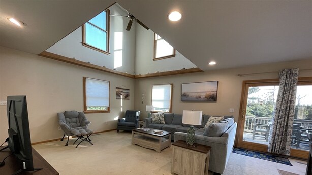 A spacious living room with a high ceiling, large windows, a television, a grey sofa, a coffee table, and a dining area with a view of the outdoors. - Soundside Home in Frisco is an Island Dream Retreat! (FRISCO)