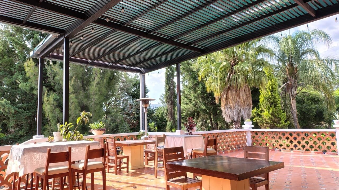 Une terrasse extérieure avec des tables et des chaises en bois, entourée d’une végétation luxuriante et surmontée d’un toit en métal.