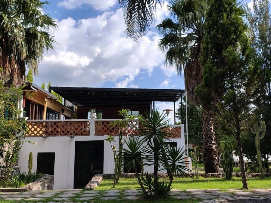 Un edificio con porche cubierto, pared de piedra y jardín con palmeras y cactus.