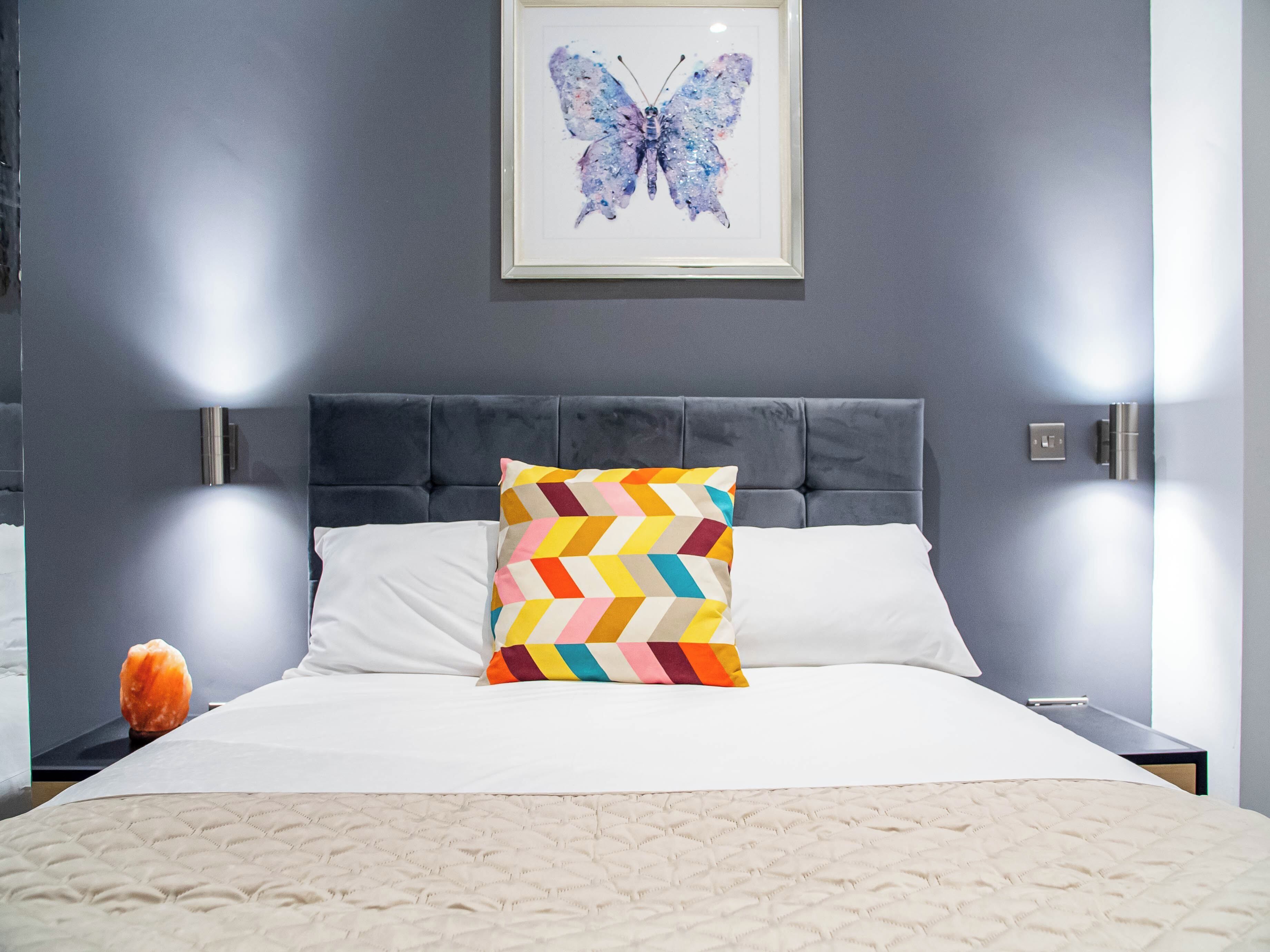 Un lit bien fait, agrémenté d’un coussin à motifs, une œuvre d’art représentant un papillon encadrée au mur et une tête de lit moderne.