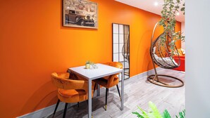 Et moderne spiseområde med et hvidt bord og orange stole, en hængestol og et billede i ramme på væggen.