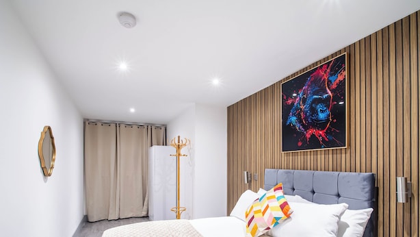 A modern bedroom with a large bed, a colorful abstract painting, a wooden panel wall, and a ceiling-mounted security camera.