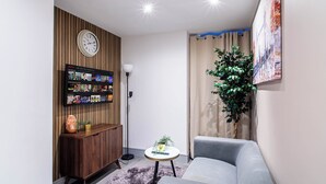 A modern living room with a grey sofa, a wooden cabinet with a clock, a wall-mounted TV displaying Netflix, a potted plant, and a painting.