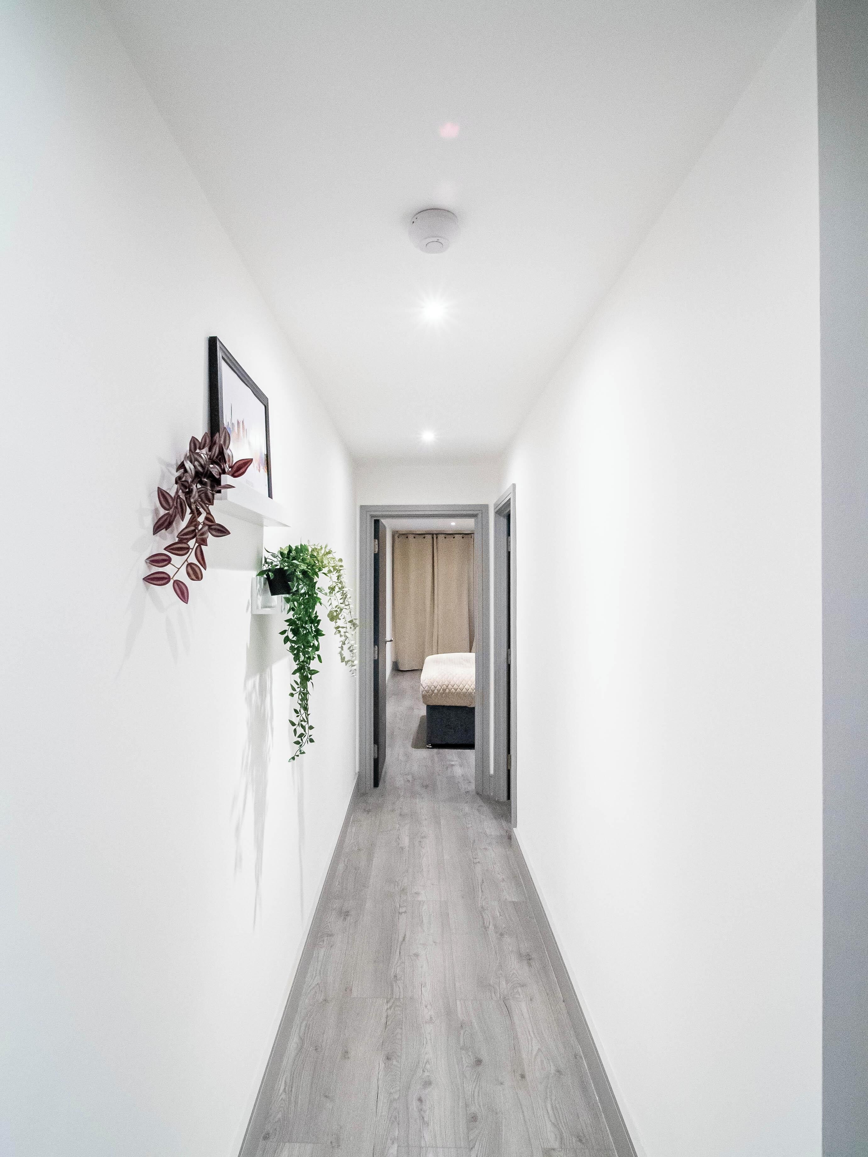 Un pasillo con piso de madera, paredes blancas y una puerta que da a una habitación con cama.