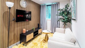Una sala de estar moderna con un panel de madera, un televisor con 'NETFLIX', un reloj, una ventana con cortinas, un sofá blanco, una mesita redonda pequeña y una planta en maceta.