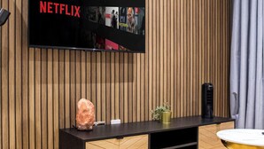 Una sala moderna con pared de paneles de madera, un reloj de pared, un televisor de pantalla plana mostrando 'NETFLIX', un mueble de entretenimiento de madera y una alfombra amarilla y suave.