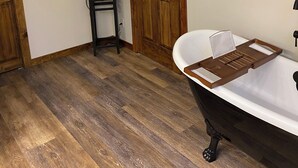 A bathroom with a wooden floor, a black bathtub, a wooden door, a wall-mounted TV, and a towel rack.
