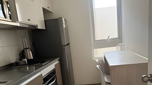 A compact kitchen with white cabinets, a stainless steel refrigerator, a dishwasher, and a window providing natural light. - Apartamento Familiar en Santiago (Santiago)