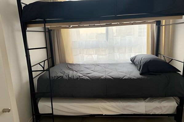 Una cama litera con colchón y almohadas grises, una ventana con cortinas y vistas de edificios.