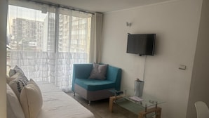 A hotel room with a bed, a blue sofa, a glass coffee table, a TV mounted on the wall, and a view of buildings through the window. - Apartamento Familiar en Santiago (Santiago)