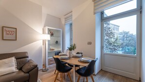 A modern living room with a brown leather sofa, a dining table set for four, and a large window offering a view of a building and trees. - Peaceful & Cozy Living in Vyšehrad with a Balcony by EasyBNB Premium (Prague)
