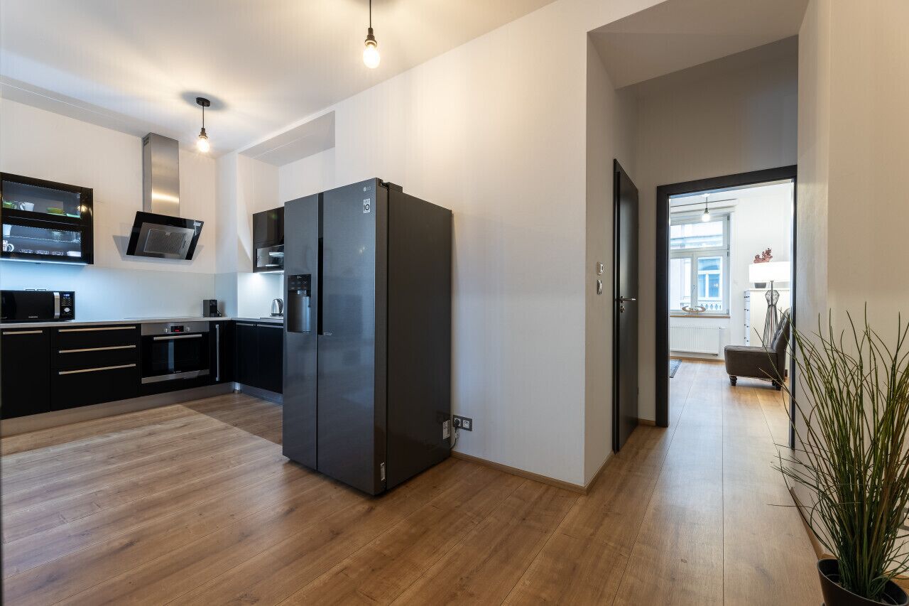 A modern kitchen with a large black refrigerator, wooden flooring, and a view into the living area.