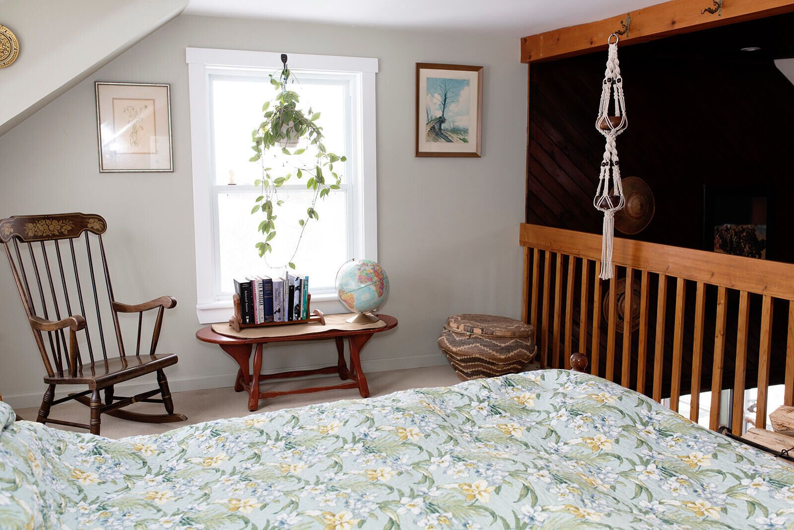 Um quarto com uma cama, uma cadeira de balanço, uma pequena mesa de madeira com livros e um globo, um macramê pendurado e uma janela com uma planta.