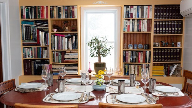 Comedor con mesa de madera dispuesta para seis personas, rodeado de estanterías llenas de libros y objetos decorativos.