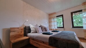 A bedroom with a wooden bed frame, multiple pillows, a bedside table, and two windows with curtains.