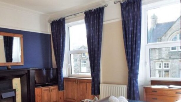A hotel room with a bed, a fireplace, wooden cabinets, a window with curtains, and a view of buildings outside. - OYO The Board Inn (Hawes)