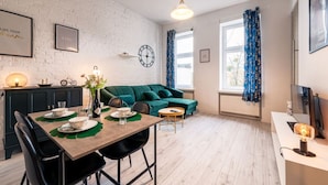 A modern living room with a green sofa, a dining table set with plates and cutlery, a clock on the wall, and a window with blue curtains. - Środmieście Apt - Top Location, Tram&Park nearby - by Rentujemy (Wroclaw)