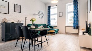 A modern living room with a dining area, featuring a green sofa, a dining table with chairs, a clock on the wall, and blue curtains. - Środmieście Apt - Top Location, Tram&Park nearby - by Rentujemy (Wroclaw)