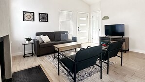Un salon moderne avec un canapé, une table basse et deux fauteuils. On y trouve un téléviseur posé sur un meuble, une petite table d’appoint avec une plante et un tapis à motifs.