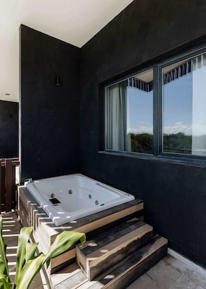A modern bathroom with a large bathtub, a window with curtains, and a view of greenery outside.