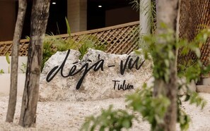En sten med teksten 'deja-vu' og 'Tulum', omgivet af grøn vegetation og et bambushegn.