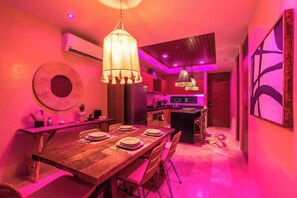 Zona pranzo con tavolo in legno apparecchiato per quattro persone, lampadario a sospensione e cucina sullo sfondo.