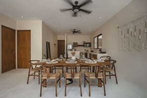 A dining room with a long table set for six, wooden chairs, ceiling fans, a kitchen in the background, and a macrame wall hanging.