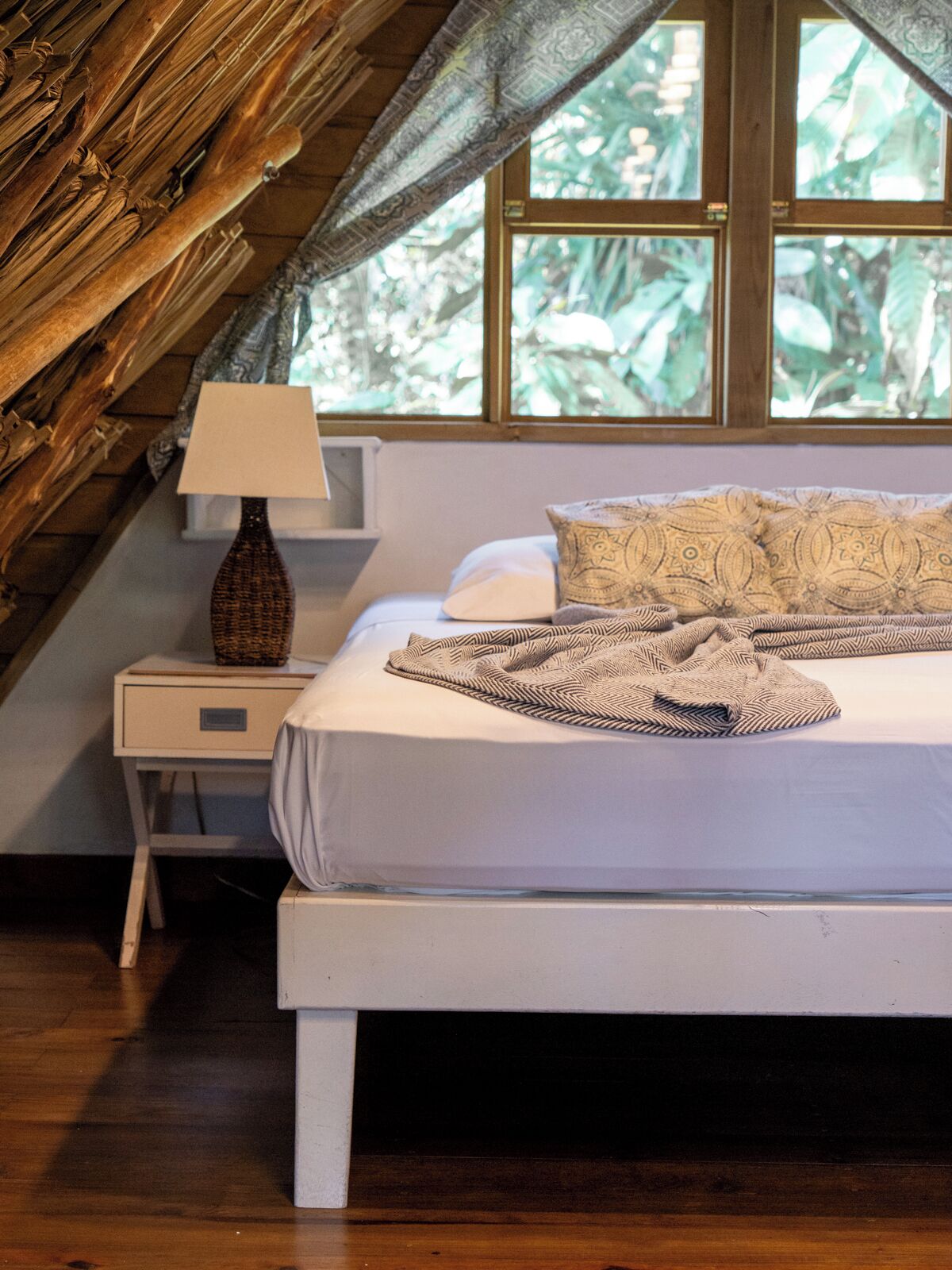 Een slaapkamer met een bed, een nachtkastje, een lamp en een raam met uitzicht op groen.