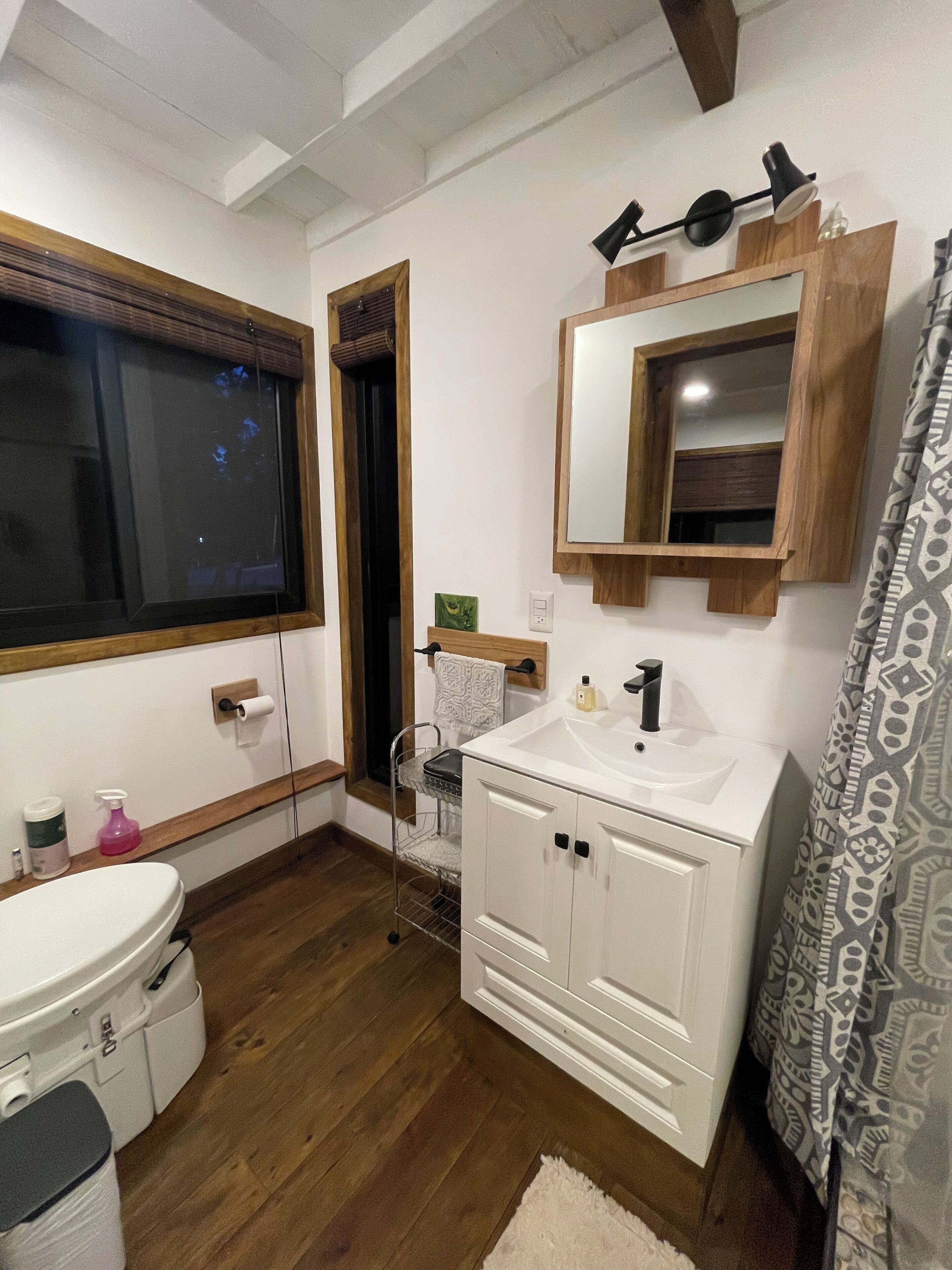 Un baño con piso de madera, gabinetes blancos, un lavamanos, un espejo y una ventana.