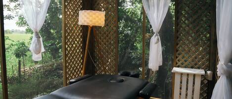 Une table de massage dans une pièce donnant sur un jardin luxuriant, avec un mur en treillis de bois et une petite chaise blanche.