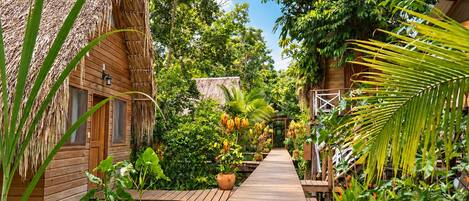 A wooden pathway leading to thatched-roof cabins surrounded by lush greenery and tropical plants.