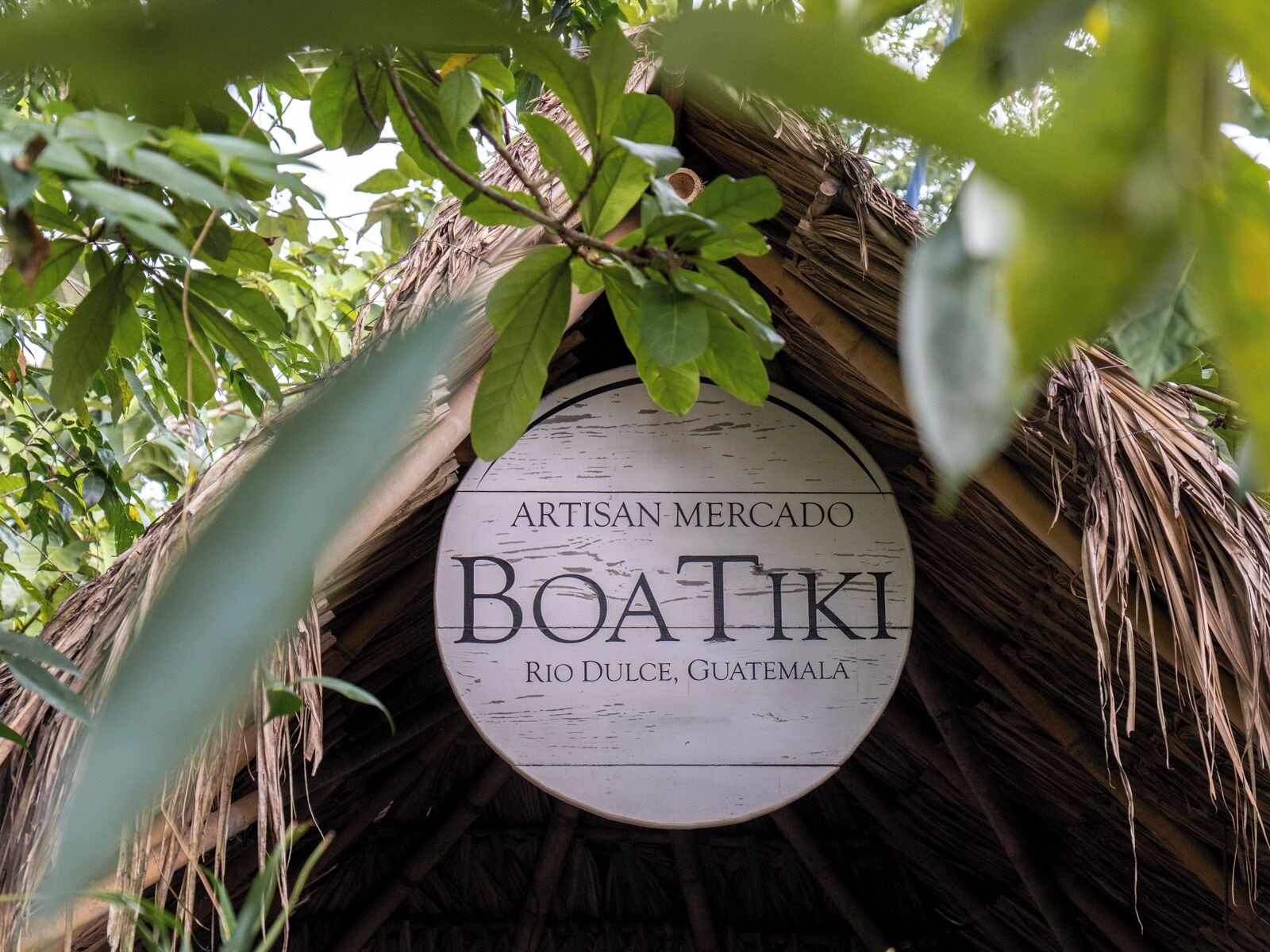 Un letrero circular con el texto 'ARTISAN-MERCADO BOATIKI RIO DULCE, GUATEMALA' colgado de una estructura de techo de paja.
