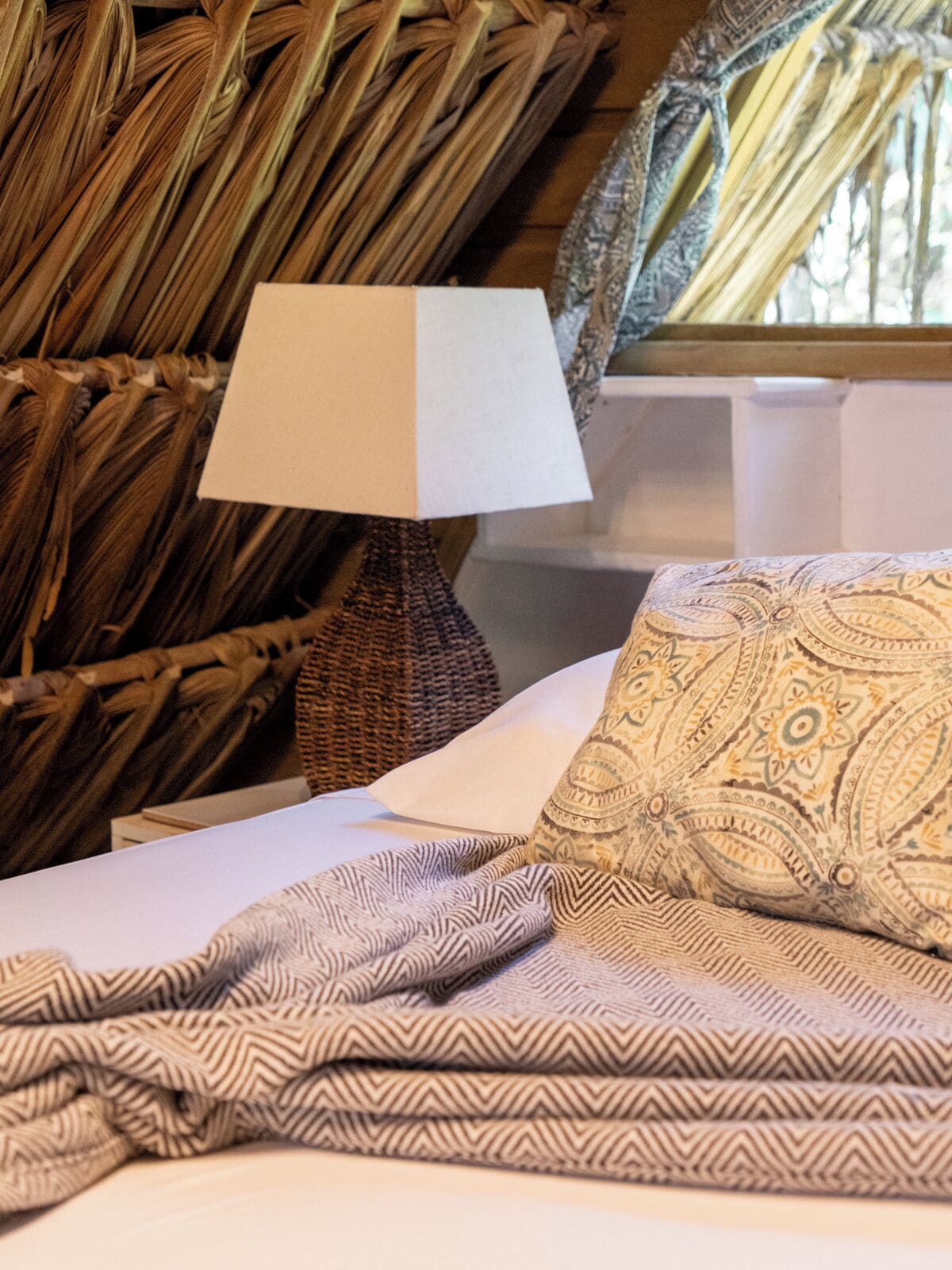 Una cama con almohadas estampadas, una colcha a rayas y una lámpara tejida.