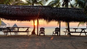 A beach scene with a thatched roof structure, two chairs, and a table under the sunset. - Boatique Hotel and Marina (Livingston)
