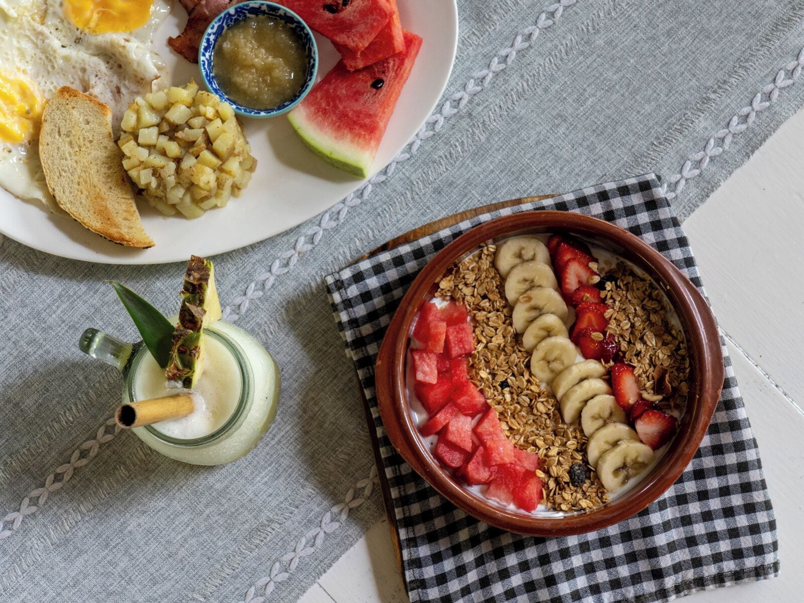 Un desayuno con un plato de huevos, tostadas, papas y sandía, junto con un tazón de yogur, frutas y granola.