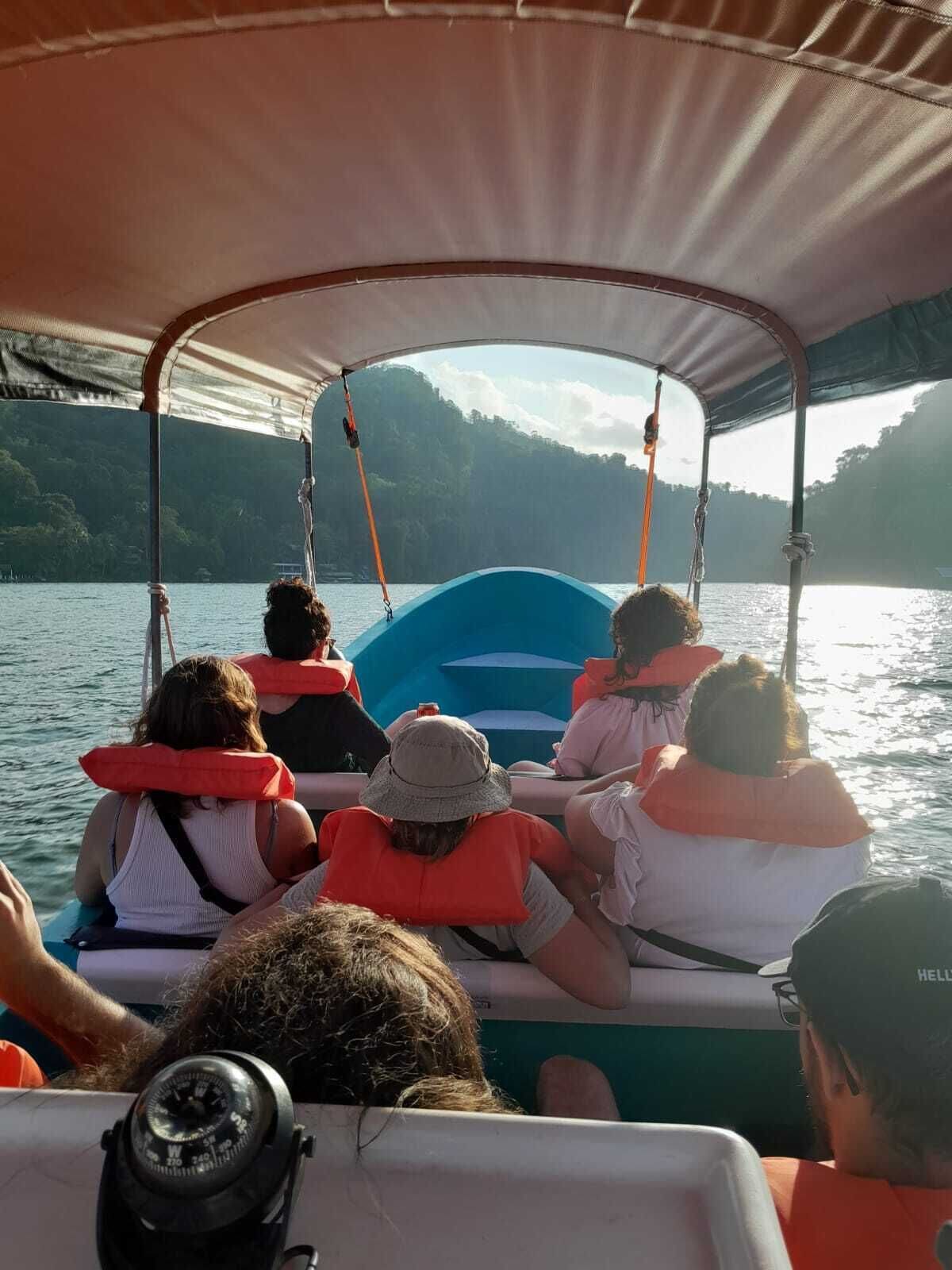 A group of people on a boat with life jackets, a canopy, and a view of a lake with trees in the background.