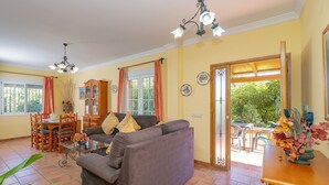 Un soggiorno con divano, tavolo da pranzo e sedie, un armadietto in legno e vista su uno spazio esterno attraverso una porta vetrata.