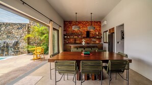 A modern dining area with a long wooden table and six chairs, a kitchen with a stone wall and wooden shelves, and a view of a waterfall and greenery outside. - Casa Wabi in Sayulita (Sayulita)