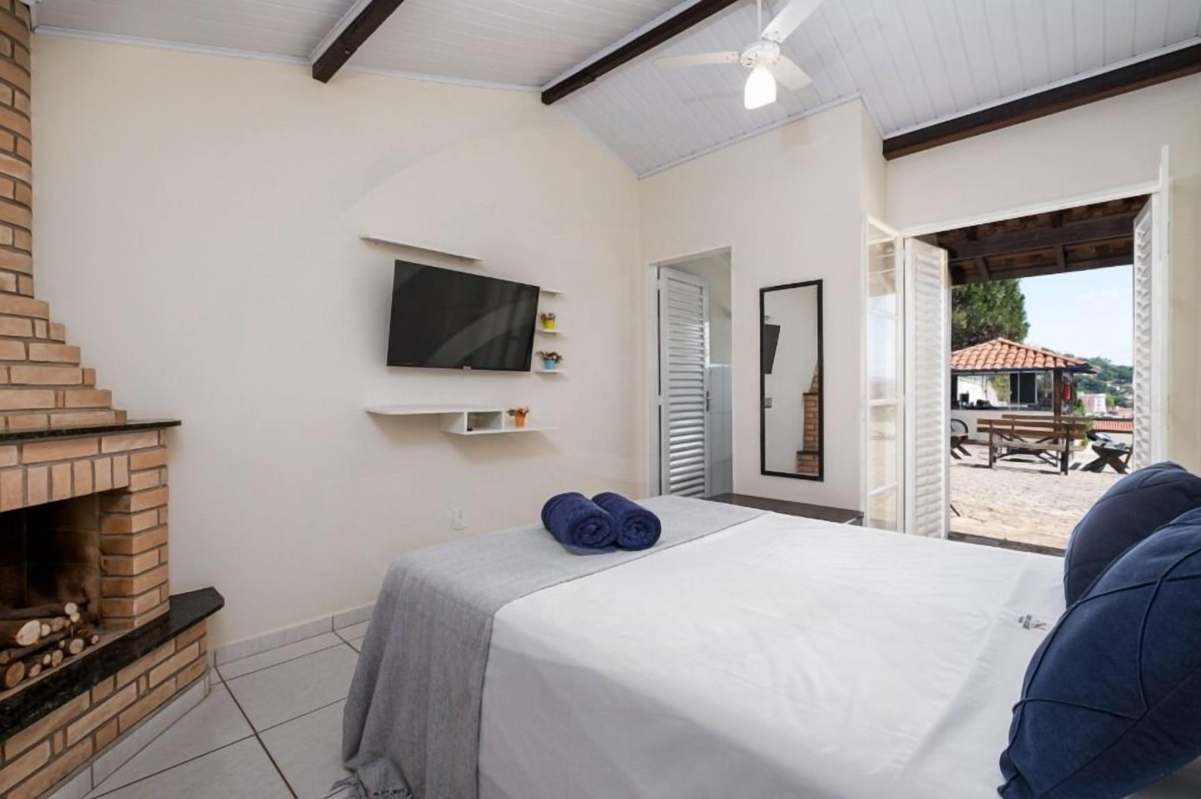 A bedroom with a brick fireplace, a bed with a white and gray bedspread, a flat-screen TV mounted on the wall, and a view of the beach through an open door.
