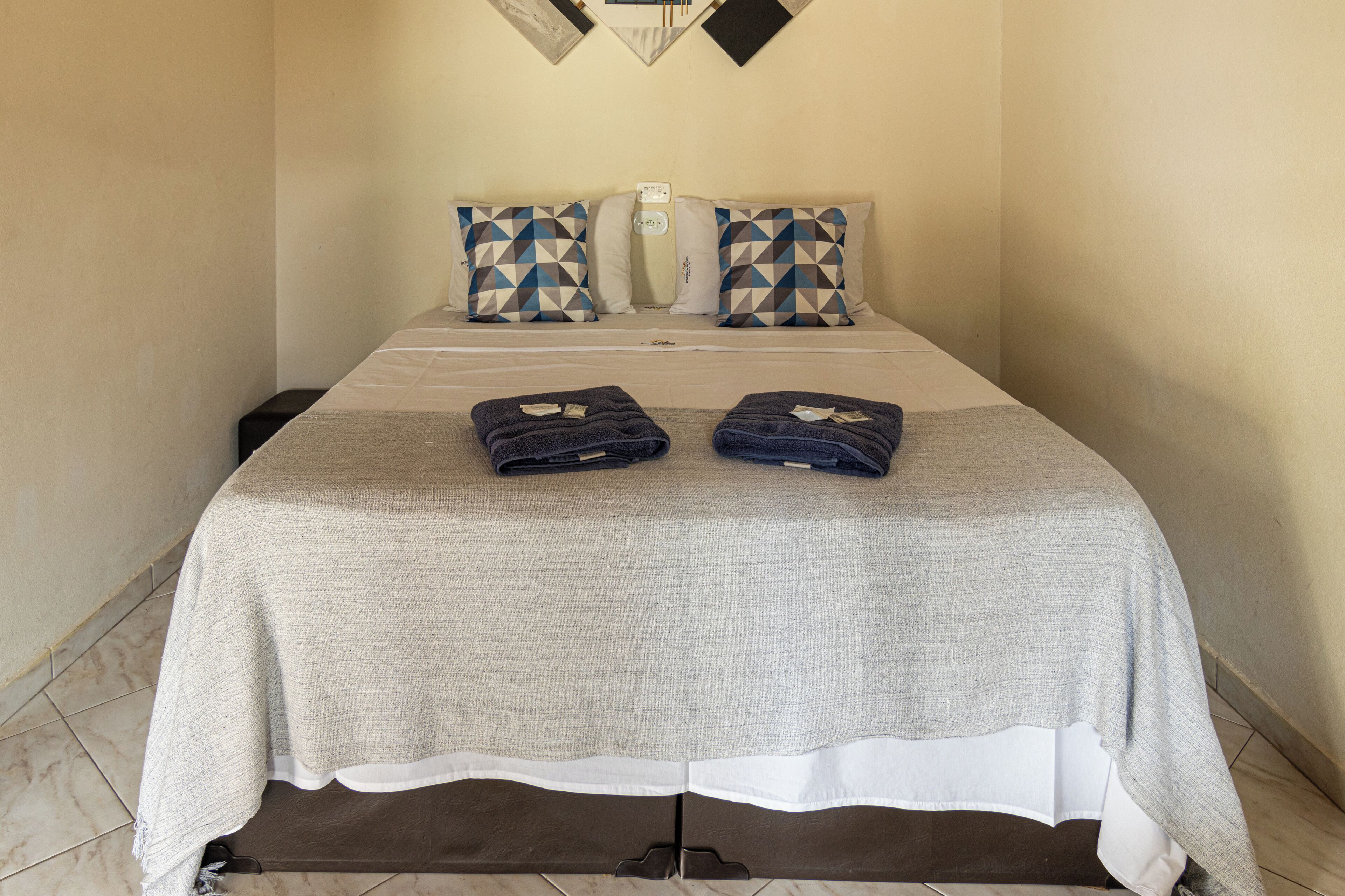 A hotel room with a bed, two pillows, a bedside table, and two folded towels.