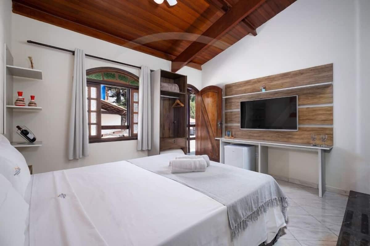 Ein Schlafzimmer mit einem Bett, einem an einer Holzwand montierten Fernseher, einem kleinen Schreibtisch und einem Fenster mit Vorhängen.