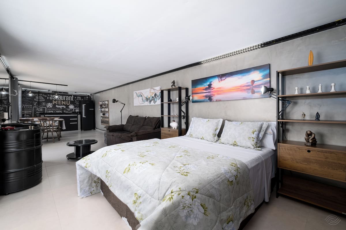 Ein modernes Schlafzimmer mit einem großen Bett, einem Schreibtisch und einem Stuhl. An der Wand hängt ein Gemälde und ein Regal zeigt dekorative Gegenstände.