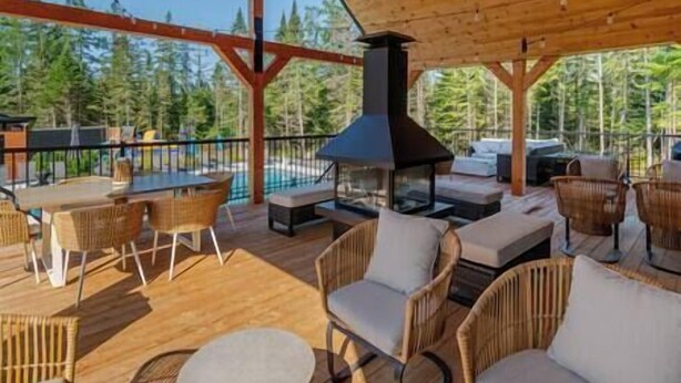 A covered outdoor seating area with wicker furniture, a central wood-burning stove, and a view of trees.