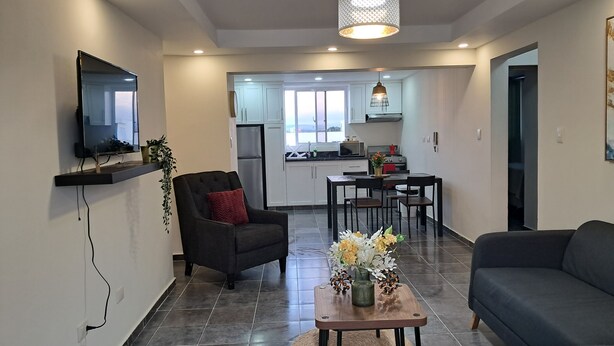 Una sala de estar moderna con un televisor de pantalla plana, un sillón oscuro, un sofá, una mesa de comedor con sillas y una cocina al fondo.