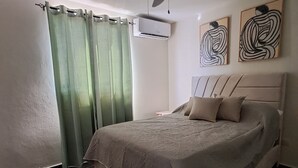 Un dormitorio con cama, ventilador de techo, equipo de aire acondicionado, cortinas verdes y arte abstracto en la pared.