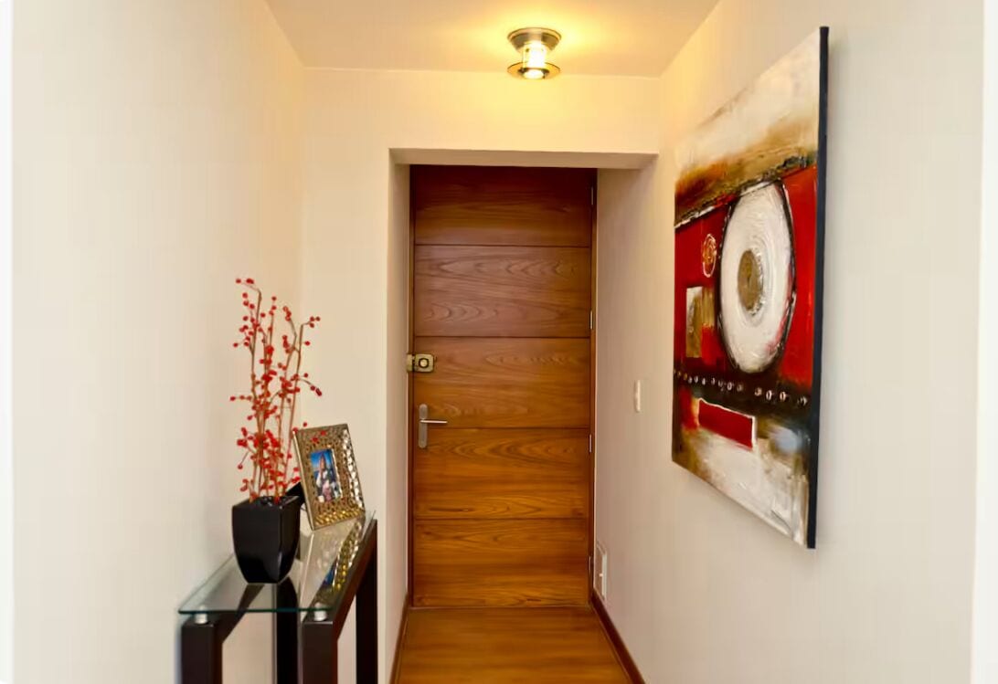 A hallway with a wooden door, a glass table with a vase and picture frame, and a large abstract painting on the wall.