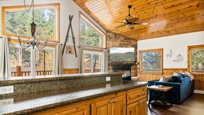 A spacious living area with a stone fireplace, wooden ceiling, and large windows offering a view of the outdoors.