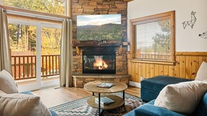 A cozy living room with a stone fireplace, a television, and a balcony with a view of trees.